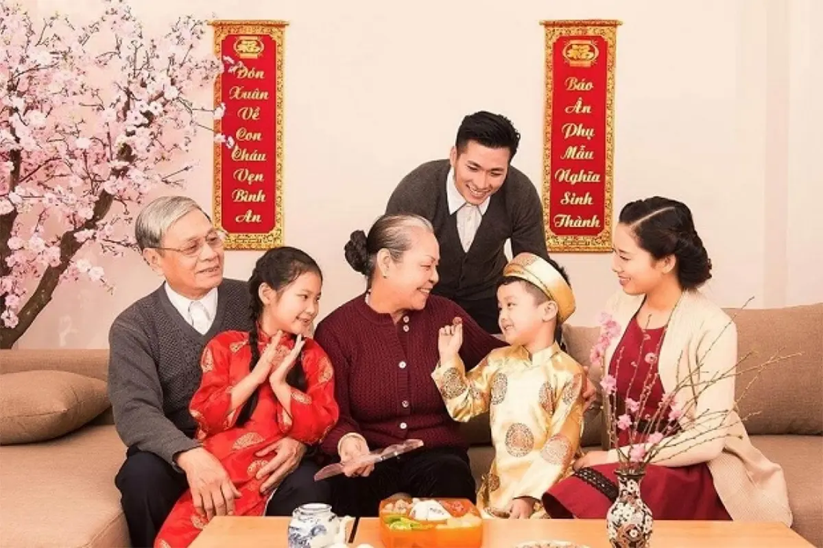 A Vietnamese family in traditional clothes celebrating Lunar New Year together at home, with red couplets and peach blossoms in the background.