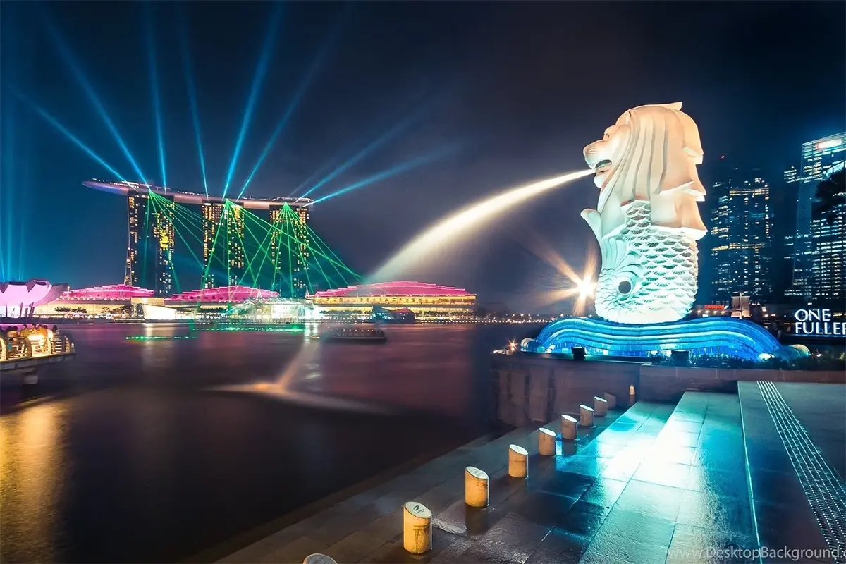 Merlion Park illuminated at night with the Marina Bay skyline and colorful lights in the background.